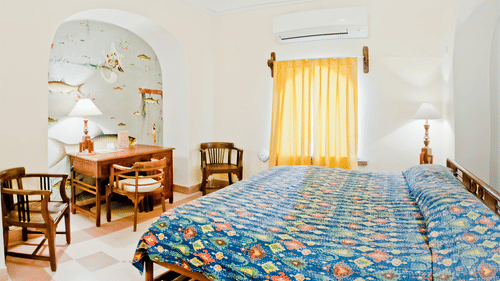 A guest room at Tijara Fort-Palace - 19th Century, Alwar features a bed with patterned linens, bedside tables with lamps, a desk with chairs in an arched alcove with a mural, a window with curtains, and an air conditioning unit.