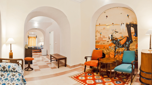 A guest suite at Tijara Fort-Palace - 19th Century, Alwar includes a bed with patterned linens, a floor lamp, and arched doorways leading to a tiled hallway with a bench and a seating area with chairs, a small table, a rug, and a mosaic wall.