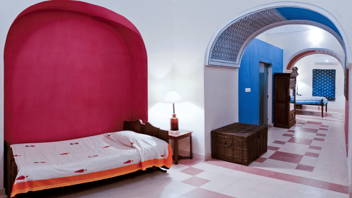 A guest room at Tijara Fort-Palace - 19th Century, Alwar features a bed with patterned linens and a red arched alcove, a bedside table with a lamp, and a view into a tiled hallway with another bed and a wardrobe.