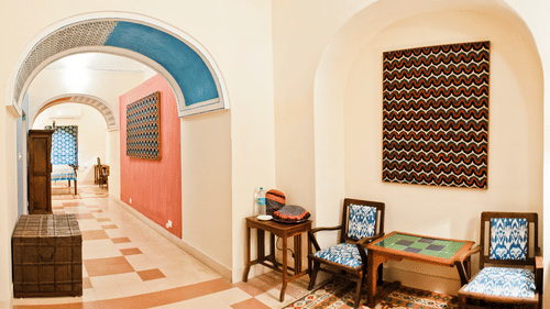 A common area at Tijara Fort-Palace - 19th Century, Alwar features arched doorways with blue accents, tiled flooring, a woven chest, artwork on the walls, chairs with patterned cushions, and small tables.