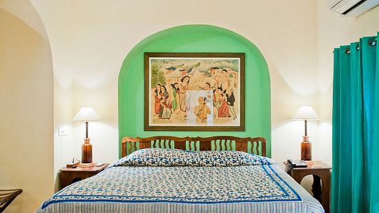 A guest room at Tijara Fort-Palace - 19th Century, Alwar features a bed with patterned linens, bedside tables with lamps, artwork in an arched alcove, a window with curtains, and an air conditioning unit.