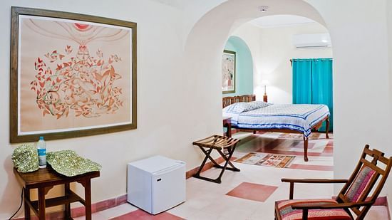 A guest suite at Tijara Fort-Palace - 19th Century, Alwar includes artwork on the wall, a table with items, a small refrigerator, a chair, and an arched opening leading to a bedroom with a bed and curtains.