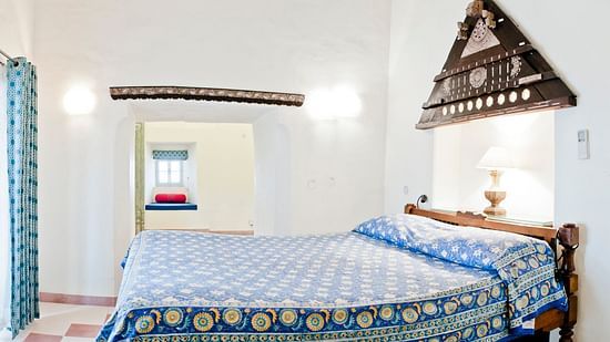 A guest room at Tijara Fort-Palace - 19th Century, Alwar features a bed with patterned linens, a bedside lamp, decorative wall hanging, a window with curtains, and a glimpse into another room.