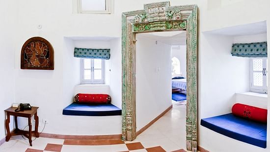 A common area at Tijara Fort-Palace - 19th Century, Alwar features tiled flooring, window seats with cushions, a small table, artwork on the wall, and an ornate doorway leading to a room with a bed.
