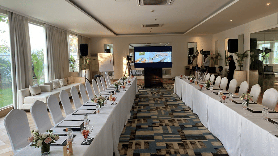 Long indoor conference room at The Mansion House resort set in U-shape with white tables and chairs, notepads, red drinks, floral arrangements, and TV presentation.