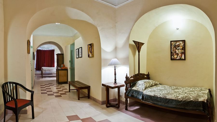 A hallway at Neemrana's Tijara Fort Palace features arched openings, tiled flooring, a floor lamp, a table lamp, a bench, a chair, artwork, and a single bed in an alcove.
