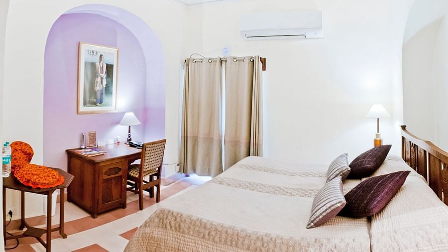 A guest room at Tijara Fort-Palace - 19th Century, Alwar features a bed with multiple pillows, bedside tables with lamps, a desk with a chair, artwork in an arched alcove, a window with curtains, and an air conditioning unit.