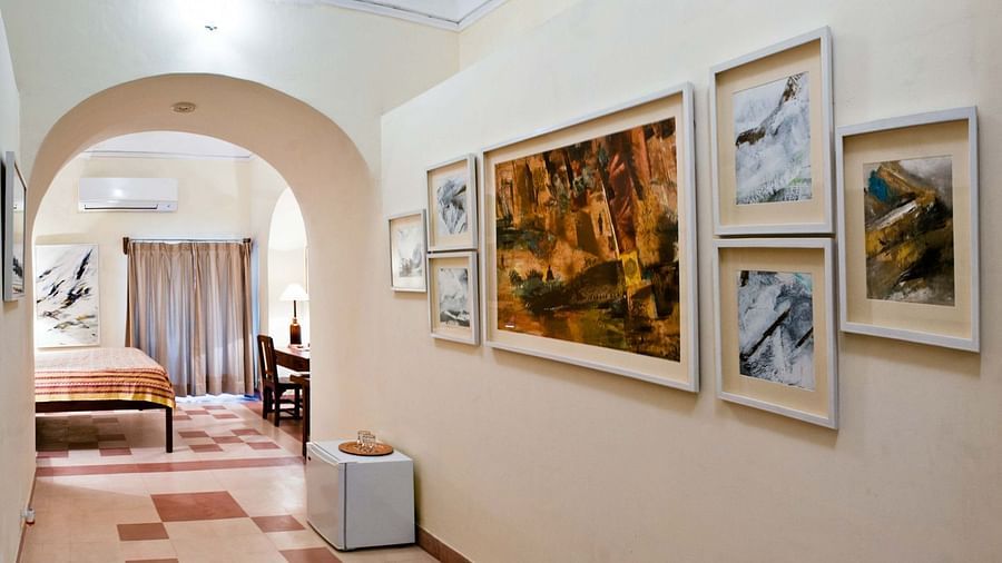 A hallway at Tijara Fort-Palace - 19th Century, Alwar features arched doorways, tiled flooring, a mini-fridge, and a collection of framed artwork on the wall. A glimpse into a guest room with a bed and desk is visible through an archway.