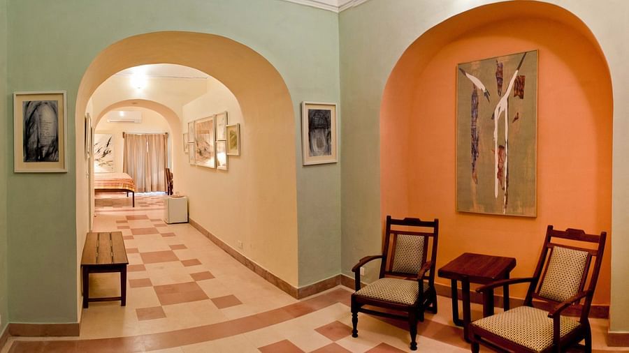 A hallway at Tijara Fort-Palace - 19th Century, Alwar features arched openings, tiled flooring, a bench, framed artwork on the walls, and a seating area with chairs and a small table in an orange arched alcove. A glimpse into a guest room is visible.