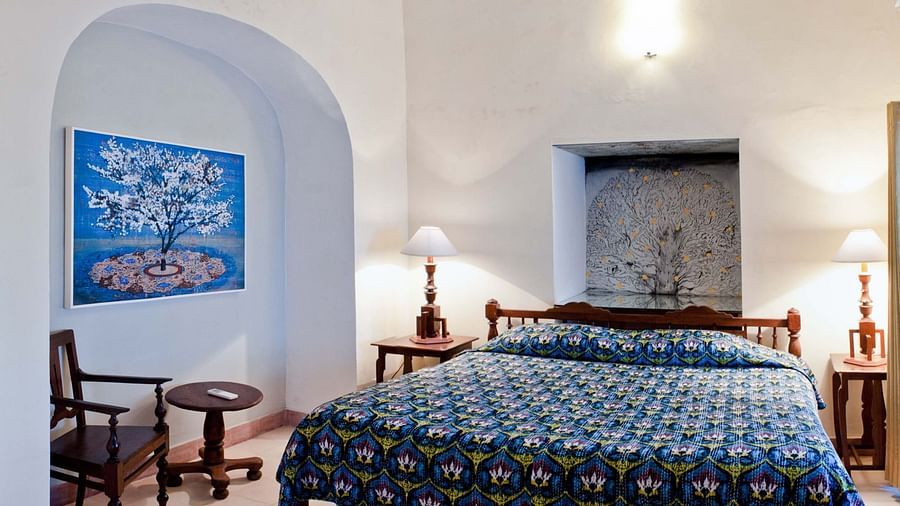 A guest room at Tijara Fort-Palace - 19th Century, Alwar features a bed with blue patterned linens, bedside tables with lamps, a chair with a small table, and blue artwork in an arched alcove.