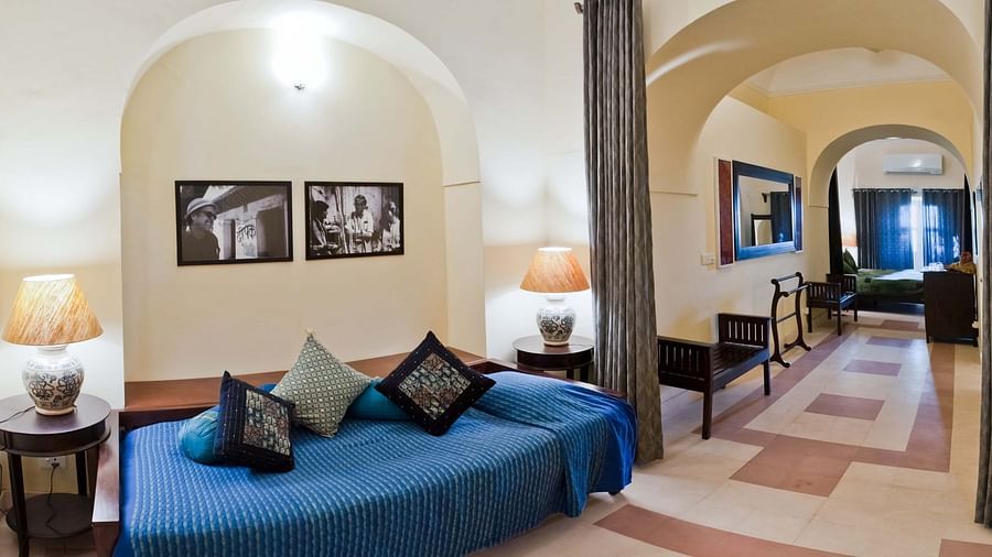 A guest room at Tijara Fort-Palace - 19th Century, Alwar features a unique curved bed with a blue cover and decorative pillows, bedside tables with lamps, framed artwork, and an arched opening leading to a tiled hallway with seating.