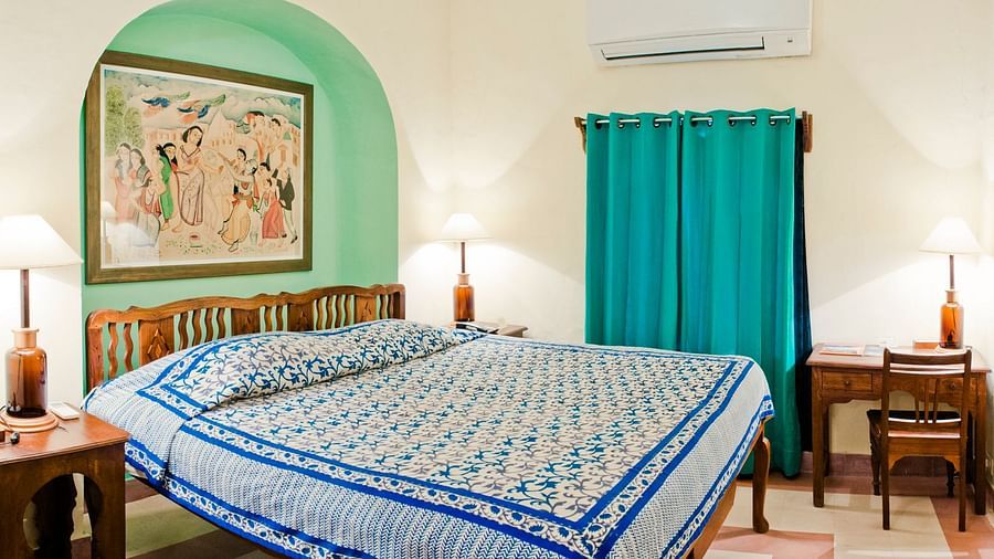 A guest room at Tijara Fort-Palace - 19th Century, Alwar features a bed with blue and white patterned linens, bedside tables with lamps, artwork on a wall with an arched alcove, a window with teal curtains, and an air conditioning unit.