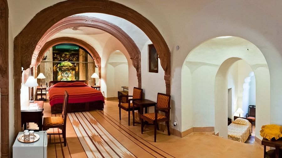 A suite at Tijara Fort-Palace - 19th Century, Alwar features arched doorways, a bedroom with a bed and bedside tables with lamps, a dining area with a table and chairs, and tiled flooring.