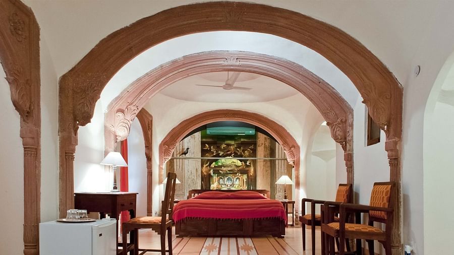 A suite at Neemrana's - Tijara Fort Palace features arched doorways, a bedroom with a bed and bedside lamps, a dining area with a table and chairs, and tiled flooring.
