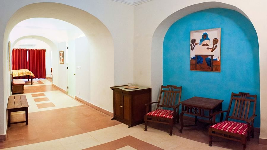 A common area at Tijara Fort-Palace - 19th Century, Alwar has arched openings, tiled flooring, chairs with striped cushions, a small table, a cabinet, and artwork in a blue arched alcove. A glimpse of a bedroom is visible.