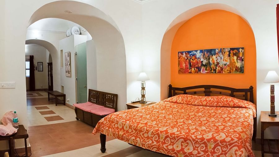 A guest room at Tijara Fort-Palace - 19th Century, Alwar includes a bed with an orange floral cover, bedside tables with lamps, artwork in an orange arched alcove, and a view into a tiled hallway with a bench.
