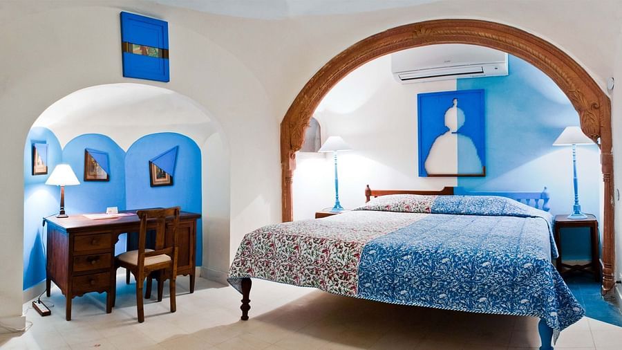 A guest room at Neemrana's Tijara Fort Palace features a bed with patterned covers, bedside tables with lamps, a desk with chairs, artwork in arched alcoves, and air conditioning.