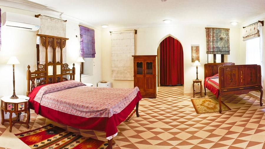 A guest room at Tijara Fort-Palace - 19th Century, Alwar features two beds with patterned covers and red accents, bedside tables with lamps, rugs, a wooden cabinet, and window coverings.