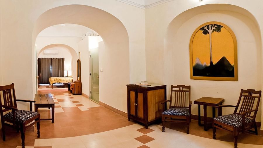 A common area at Tijara Fort-Palace - 19th Century, Alwar features arched openings, tiled flooring, chairs, small tables, a cabinet, and artwork in an arched alcove. A glimpse of a bedroom is visible.