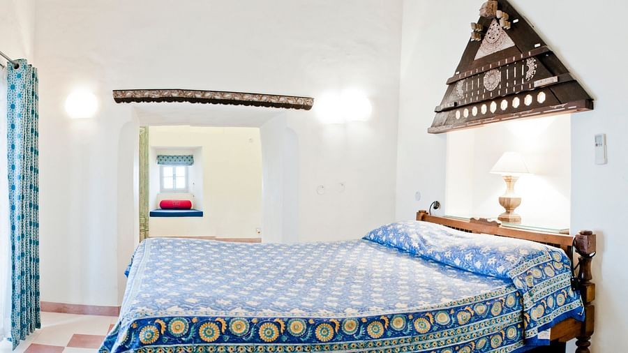 A guest room at Tijara Fort-Palace - 19th Century, Alwar features a bed with patterned linens, a bedside lamp, decorative wall hanging, a window with curtains, and a glimpse into another room.