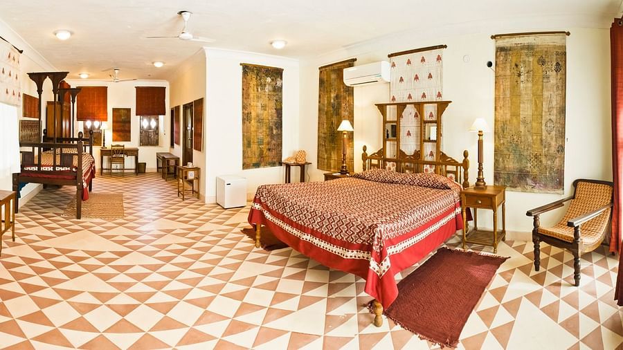 A spacious guest room at Tijara Fort-Palace - 19th Century, Alwar includes a bed with a patterned cover and red accents, bedside tables with lamps, chairs, a desk, multiple windows with coverings, artwork, and tiled flooring.