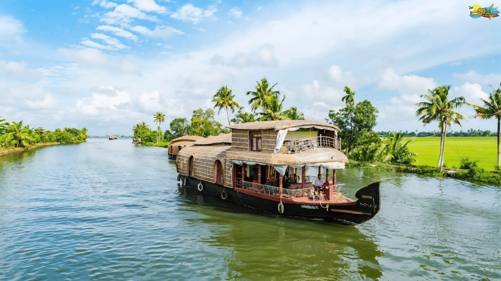 A large traditional Kerala houseboat cruising on a wide backwater canal past palm trees and paddy fields.