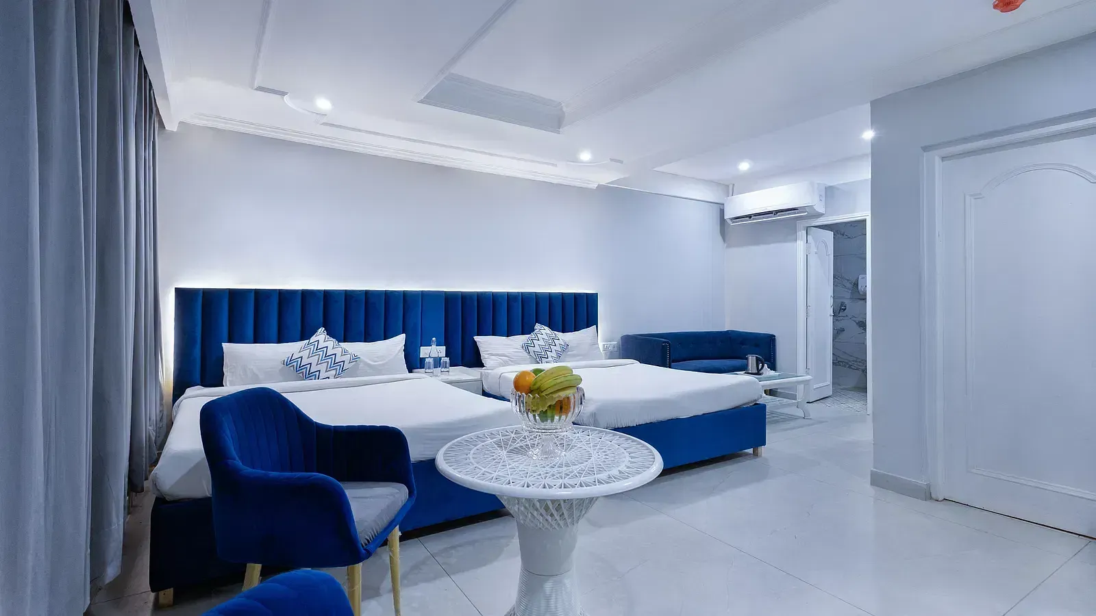 Family Room featuring blue-accented large beds, a white table with a fruit plate, and a clean, modern interior. - Hotel Downtown17, Chandigarh