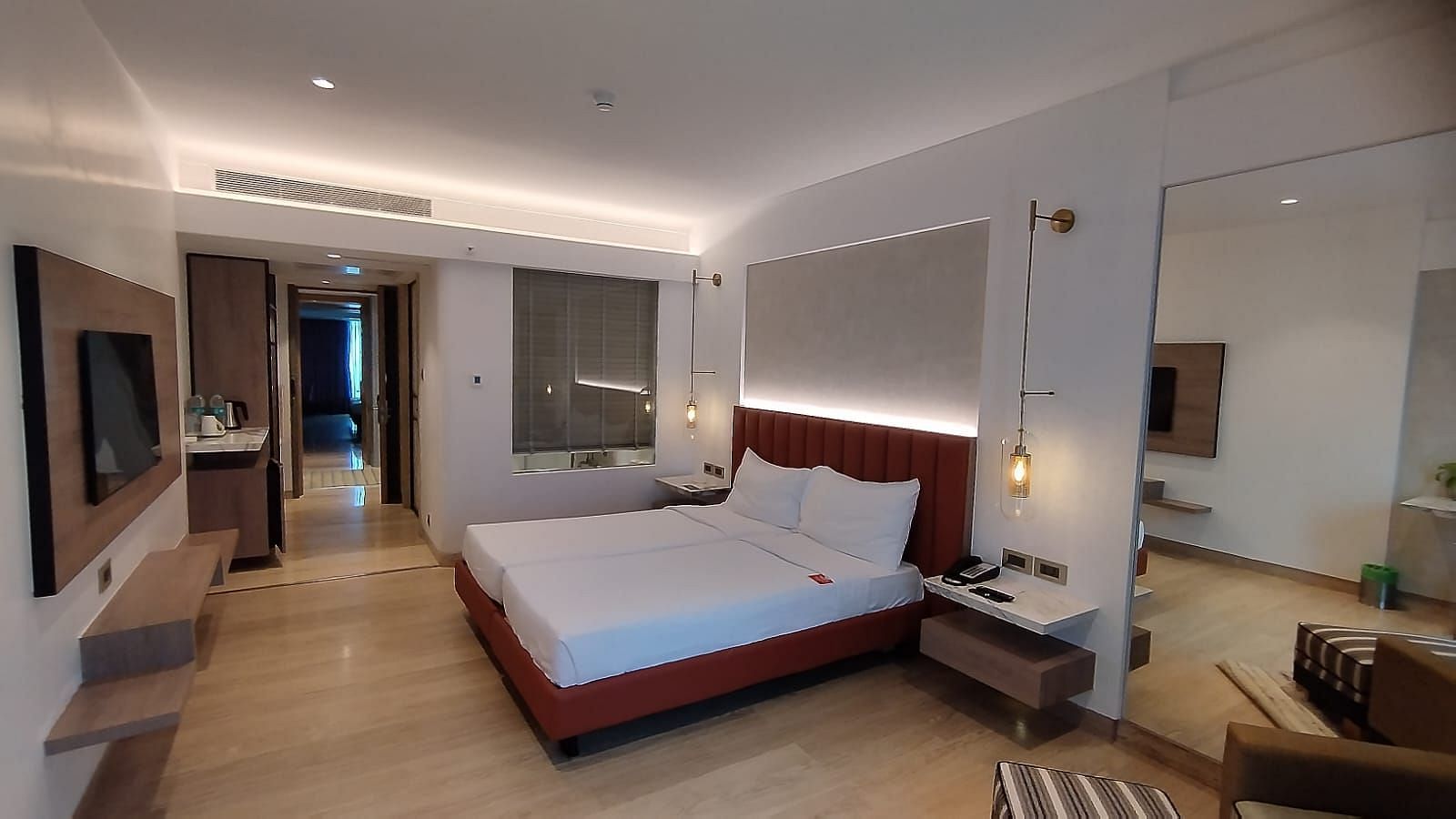 A warmly lit executive bedroom in Mirage Hotel, featuring twin beds, a large mirror, lounge chairs, and an entertainment unit.