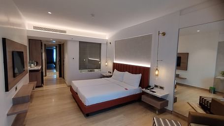 A warmly lit executive bedroom in Mirage Hotel, featuring twin beds, a large mirror, lounge chairs, and an entertainment unit.