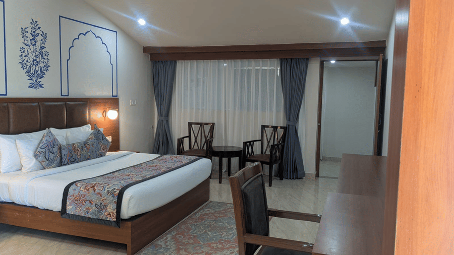 A Family Room with a cosy ambience features a neatly made bed with decorative pillows and a patterned throw. There’s a wooden desk and two chairs near the window with elegant blue curtains. Soft lighting and wall art add a sophisticated touch at Bamboo Saa