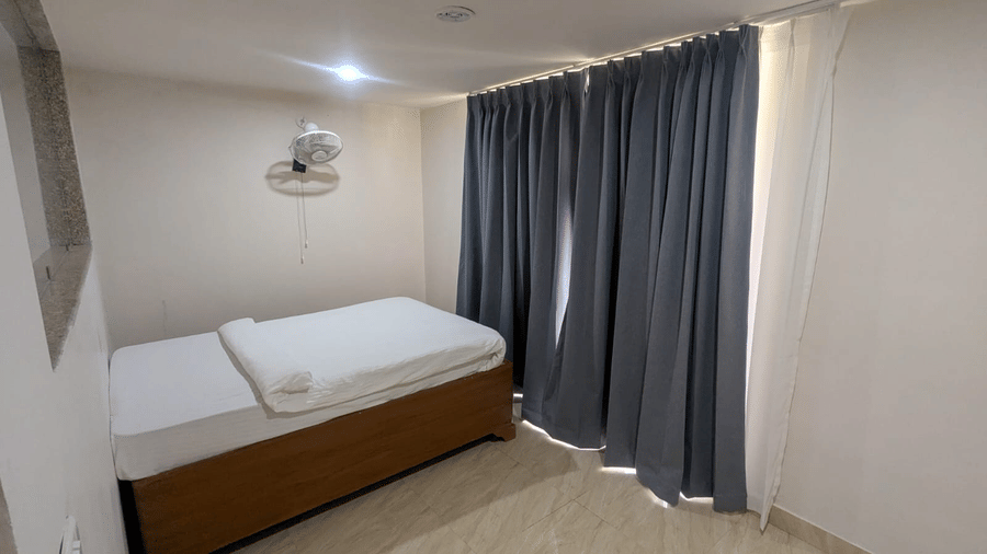A Family Room with a single bed, white bedding, and a wall-mounted fan. Dark curtains cover a large window, creating a calm, cosy atmosphere at Bamboo Saa Sunrrise Wedding & Wellness Resort, Jaipur.