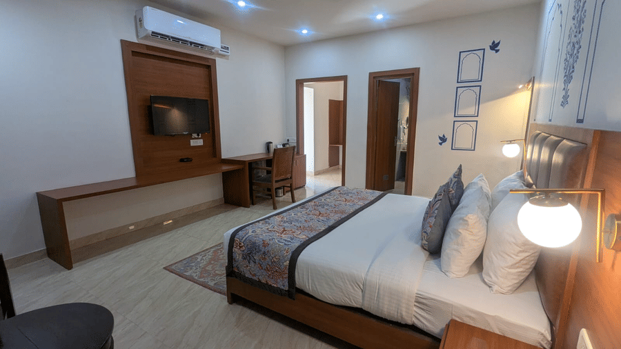 A Family Room with a neatly made bed, decorative pillows, and wall art. A desk beneath a wall-mounted TV adds functionality. Bright, calming ambiance at  Bamboo Saa Sunrrise Wedding & Wellness Resort, Jaipur.