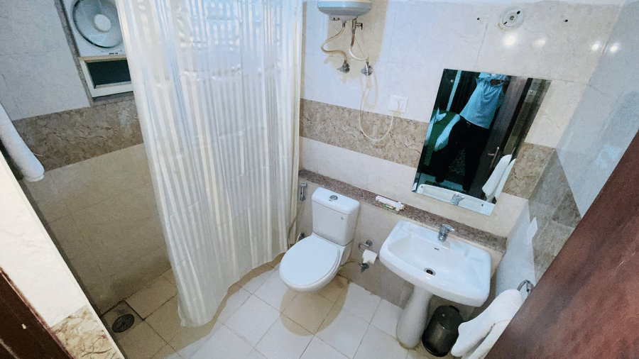 A compact bathroom with a toilet, sink, mirror, wall-mounted geyser, and a shower curtain at Livbox Rudrapur.