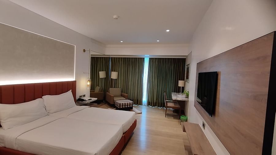An executive bedroom in Mirage Hotel, featuring twin beds, decorative head-rest, lounge chairs, an entertainment unit, and a large wall to wall window with closed drapes.