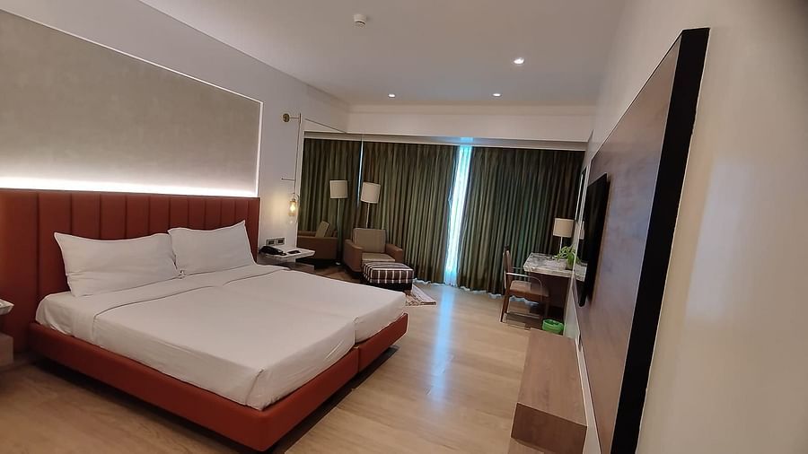 A warmly lit executive bedroom in Mirage Hotel, featuring twin beds, decorative head-rest, lounge chairs, an entertainment unit and a large wall to wall window with long drapes.