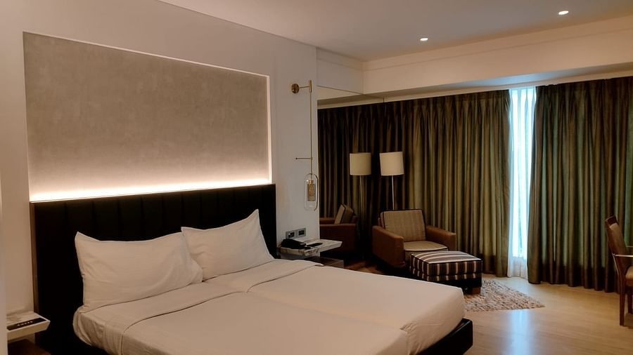 A warmly lit executive bedroom in Mirage Hotel, featuring twin beds, decorative head-rest, lounge chairs, and a large wall to wall window with closed drapes.