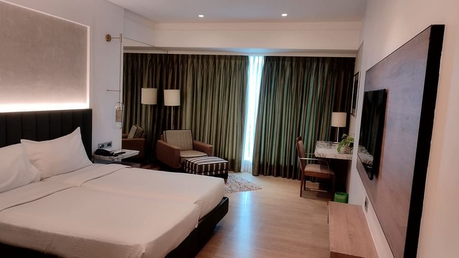 A well furnished executive bedroom in Mirage Hotel, featuring twin beds, decorative head-rest, lounge chairs, an entertainment unit and a large wall to wall window with closed drapes.