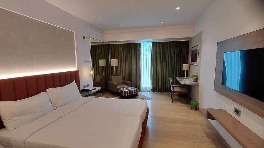 A warmly lit executive bedroom in Mirage Hotel, featuring twin beds, decorative head-rest, lounge chairs, a wooden panelled entertainment unit and a large wall to wall window with closed drapes.