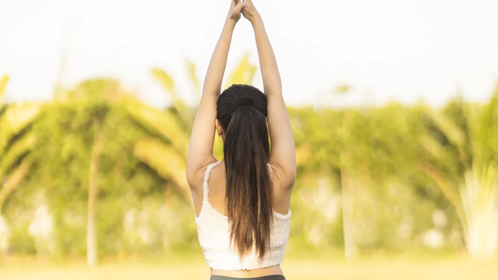 Back view of a woman wearing sports gear and in a yoga posture with blurry background of grass lawn and trees -Daksh Eden Greenz, Sasan Gir