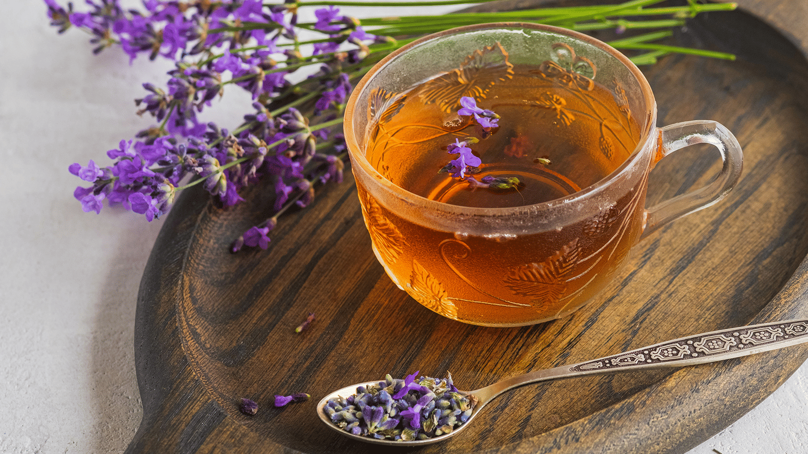 Glass cup of herbal tea with lavender sprigs and a spoon of dried flowers on a wooden board.