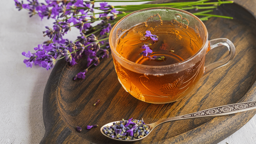 Glass cup of herbal tea with lavender sprigs and a spoon of dried flowers on a wooden board.