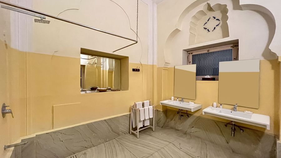 A bathroom with two sinks, a mirror, a shower head, a towel rack, a television, and an arched architectural detail at Tijara Fort-Palace - 19th Century, Alwar.