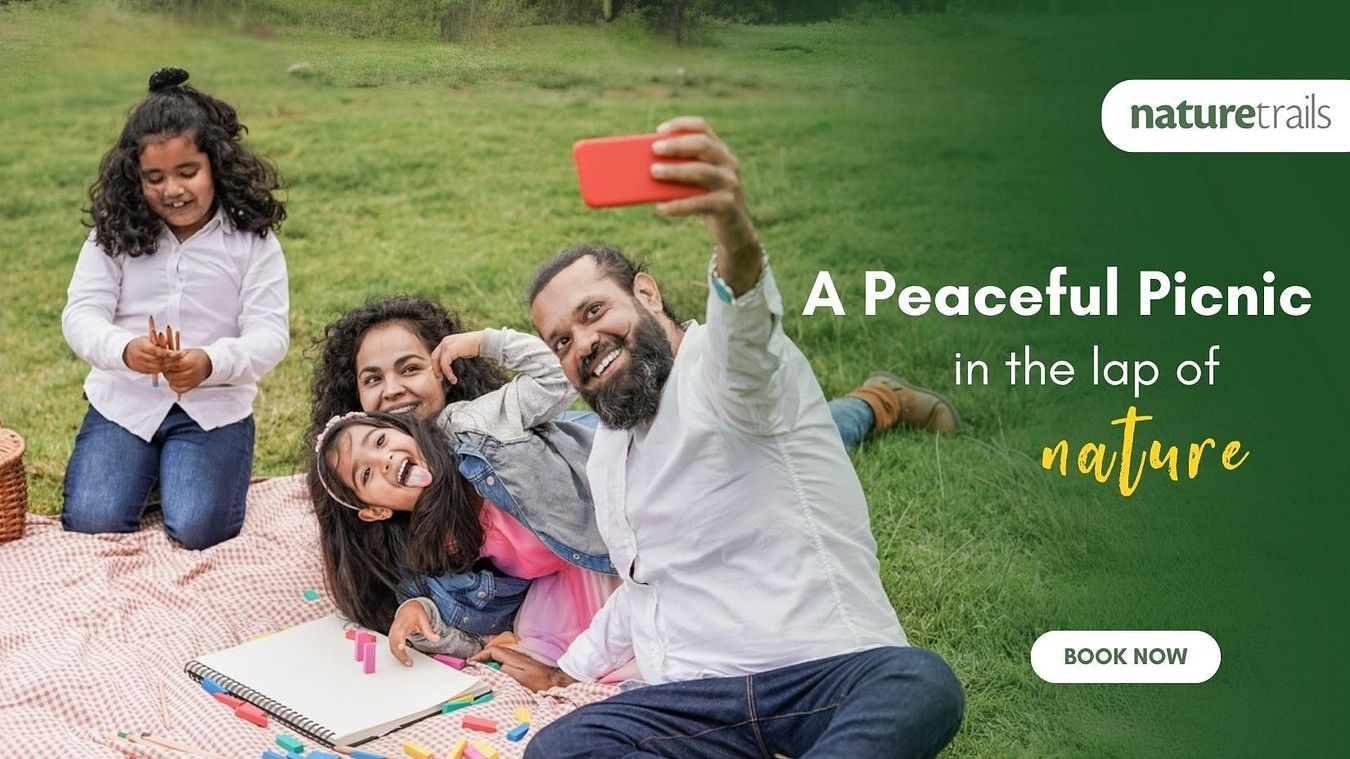 Family enjoying a picnic on a grassy field while taking a selfie. Text reads a peaceful picnic in the lap of nature. Book now.