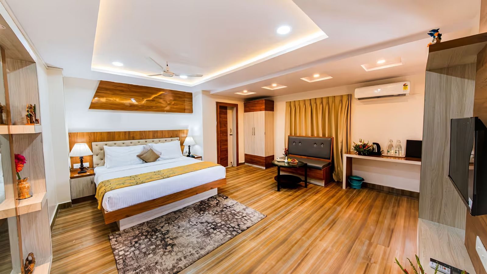  A spacious room with a bed and bedside table with lamps, a wall-mounted shelf adorned with decorative elements, and a sofa, table, and desk positioned on the opposite side.