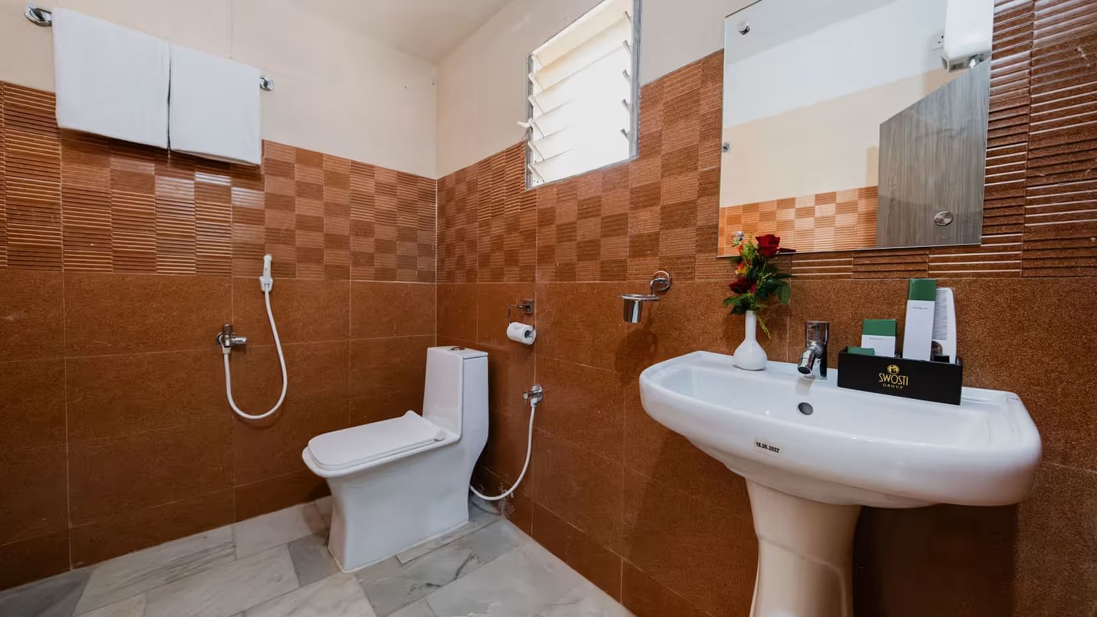 A bathroom with a western toilet, towels hung on a rack, a washbasin with toiletries on the side, and a mirror mounted directly above it.