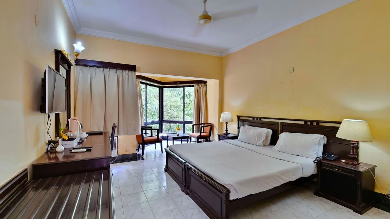  A spacious room with a large bed, a wall-mounted television opposite it, and a full-length window to the side, accompanied by wooden furniture.