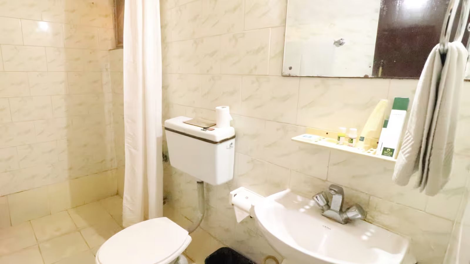 A bathroom with a western toilet, towels hung on a rack, a washbasin with toiletries on the side, and a mirror mounted directly above it.