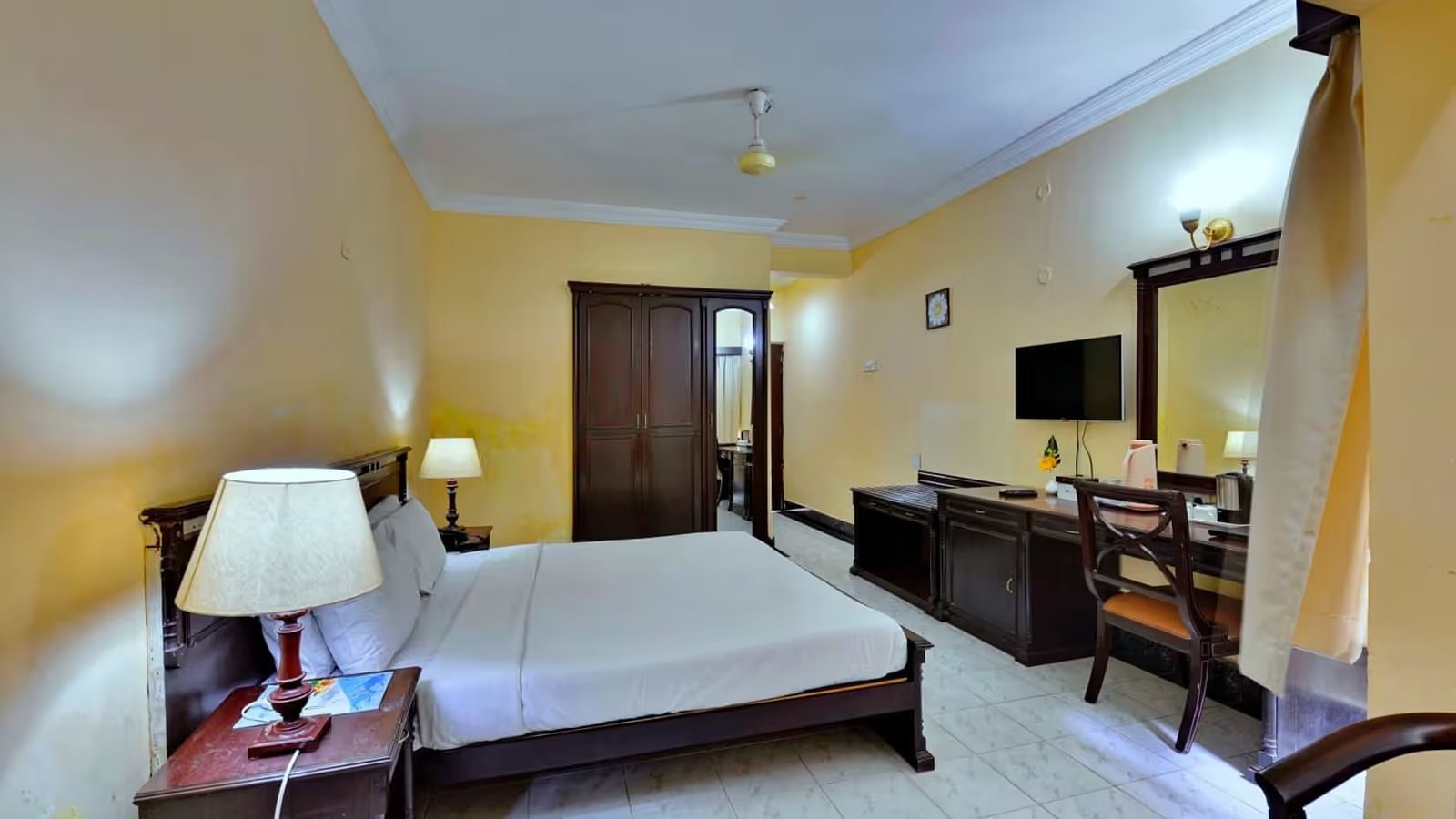  A spacious room with a bed and bedside table with lamps, a wall-mounted shelf adorned with decorative elements, and a sofa, table, and desk positioned on the opposite side.
