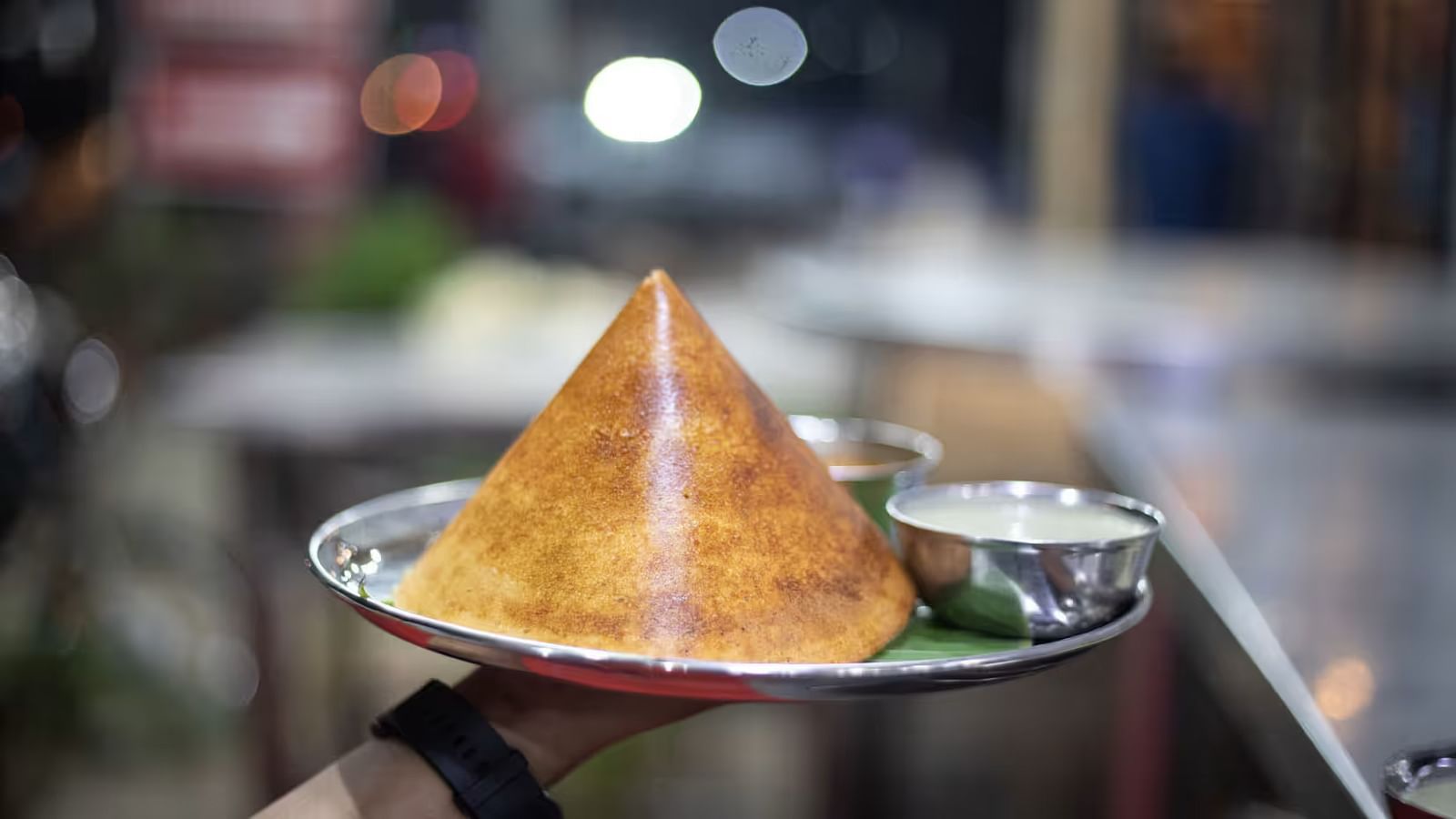  A hand holding a plate with dosa rolled into a cone, accompanied by two types of chutney in small bowls.