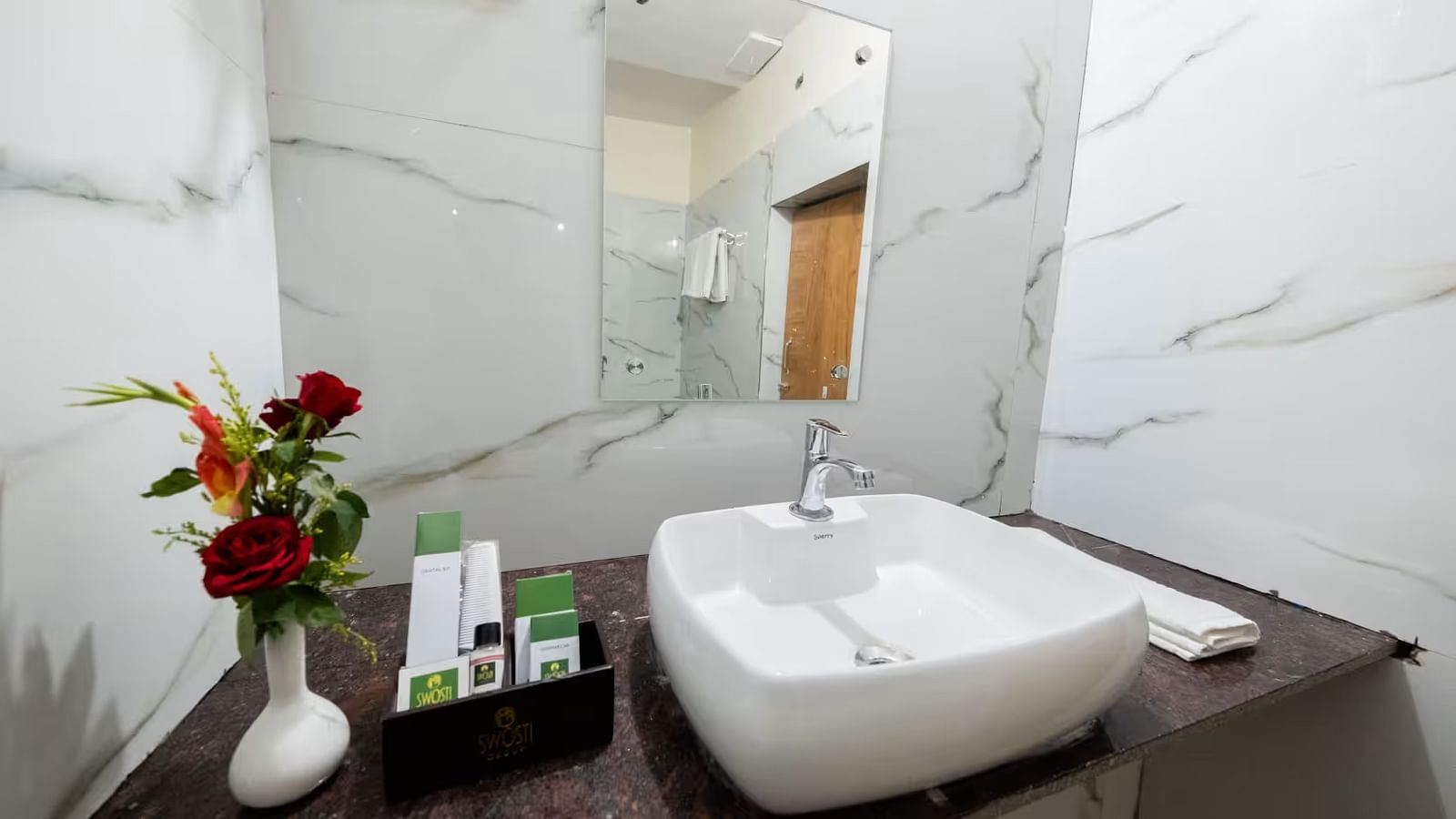  The bathroom of the Standard Room features an elegant washbasin with a mirror above it, a tray beside it with essential toiletries, and a flower vase with an assortment of flowers for decoration.